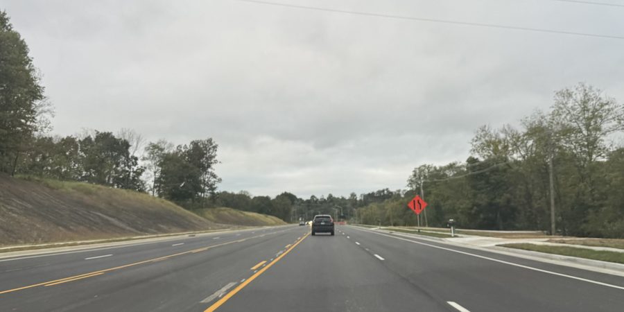 Bristow Road looking south from the driver's seat: two new lanes in each direction with a double yellow center line and fresh white lane lines, no cones, one car ahead, and an orange lane-ends sign on the right shoulder.